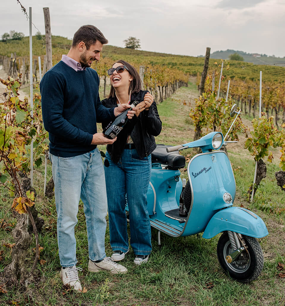 A man and a woman in a vineyard holding a bottle of wine with a blue vespa behind them
