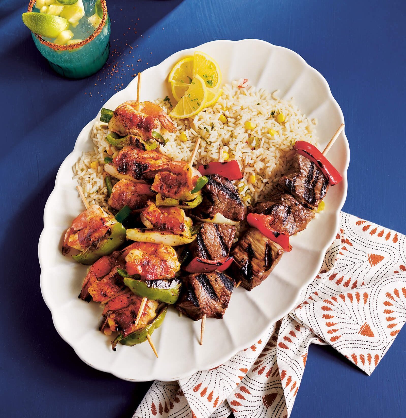 Grilled beef and shrimp skewers with drinks nearby on a bright blue surface