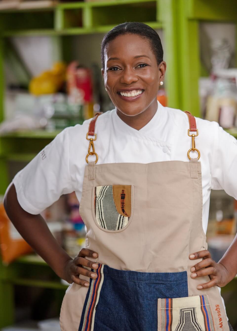 A woman, Selassie Atadika, wears a tan apron and smiles at the camera with her hands on her hips