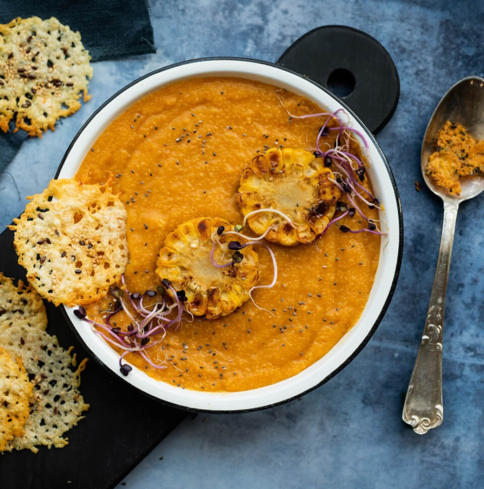 A bowl of carrot soup with crispy corn wheels on a blue tabletop with a spoon next to it