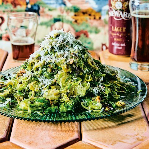 A plate of Brussels sprouts on a green glass plate with a stone tiled surface and beer visible in the background