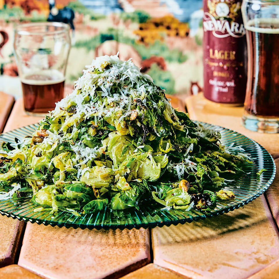 A plate of Brussels sprouts on a green glass plate with a stone tiled surface and beer visible in the background