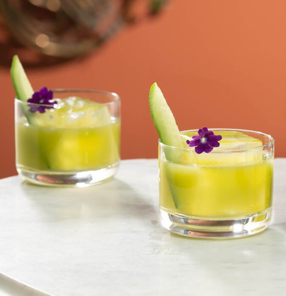 Two green cocktails in short glasses with cucumber and purple flower garnishes