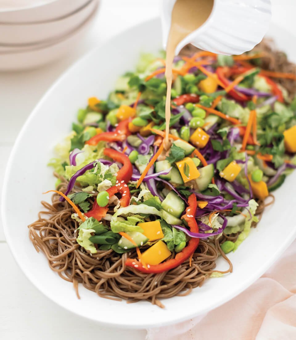 A white dish of soba noodle salad with vegetables and dressing being poured over it