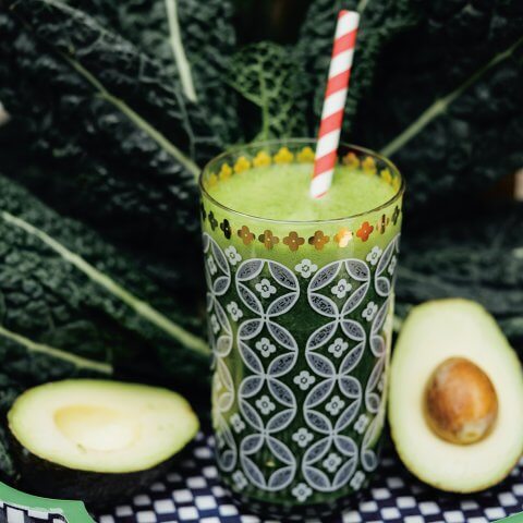 A green smoothie in a patterned glass with a sliced avocado next to it and green vegetable leaf behind
