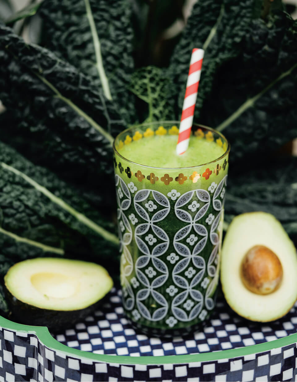 A green smoothie in a patterned glass with a sliced avocado next to it and green vegetable leaf behind