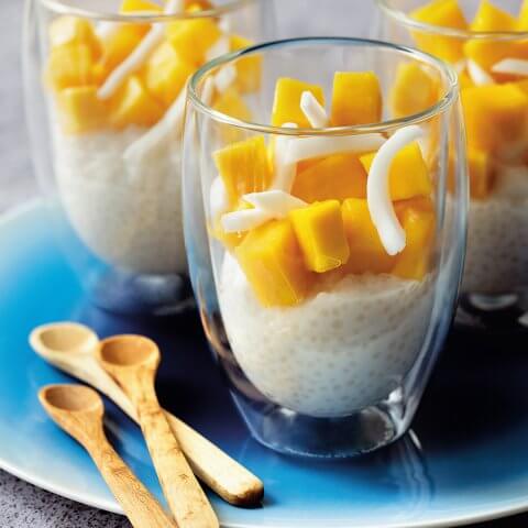 Three glasses with tapioca pudding topped with mango on a blue plate with small wooden spoons