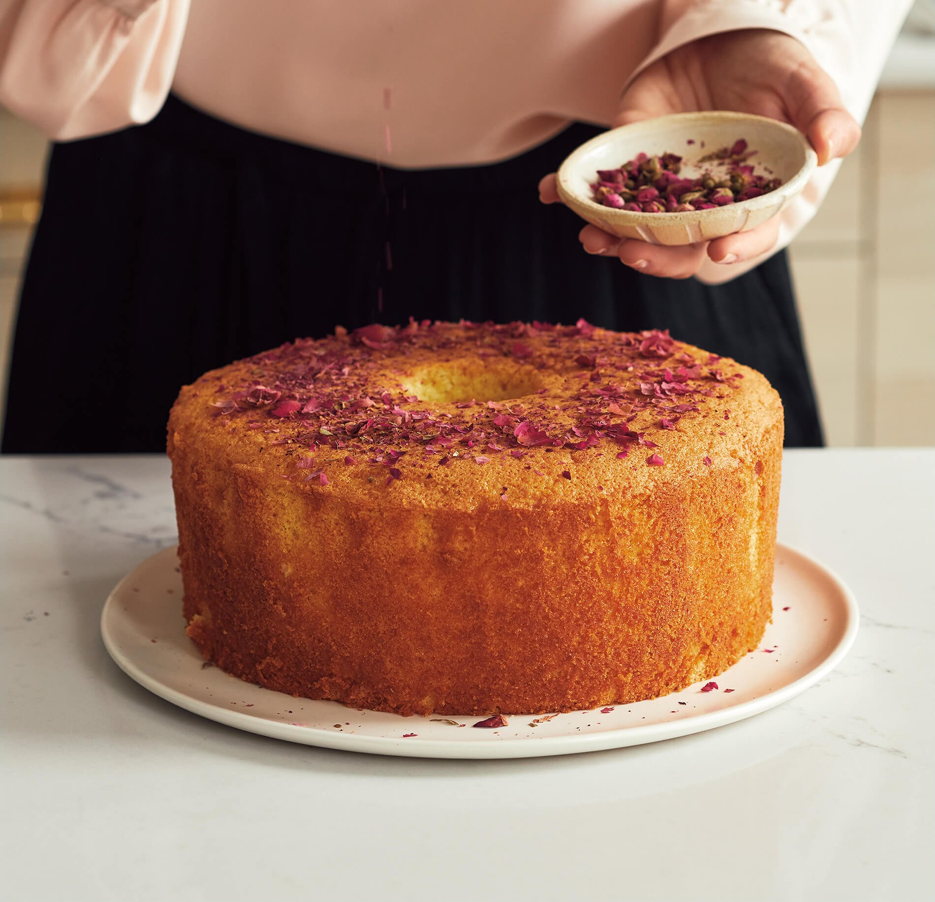 A person in a silk pink top sprinkles dried rose petals on a bundt cake over a white countertop