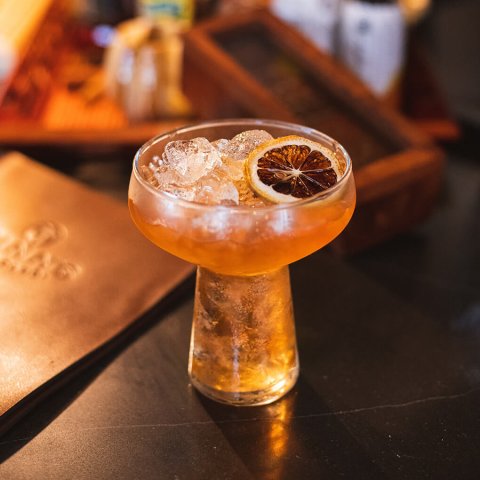 An orange-hued cocktail with a dried citrus garnish on a dark wood table