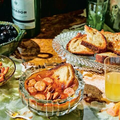 A glass bowl with chorizo and sauce, a plate with toasted bread and green bottles visible in the background