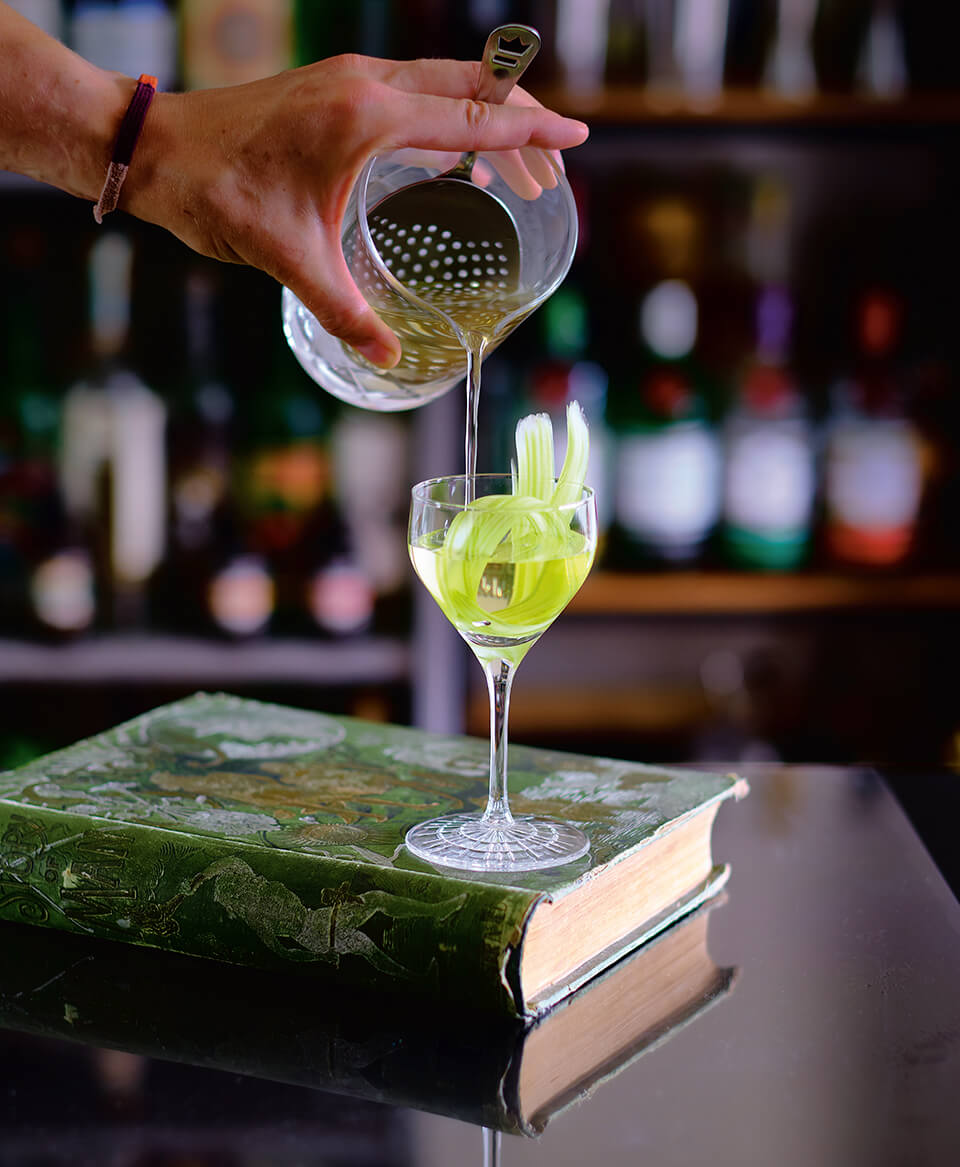 A cocktail being poured into a glass resting on top of a green book