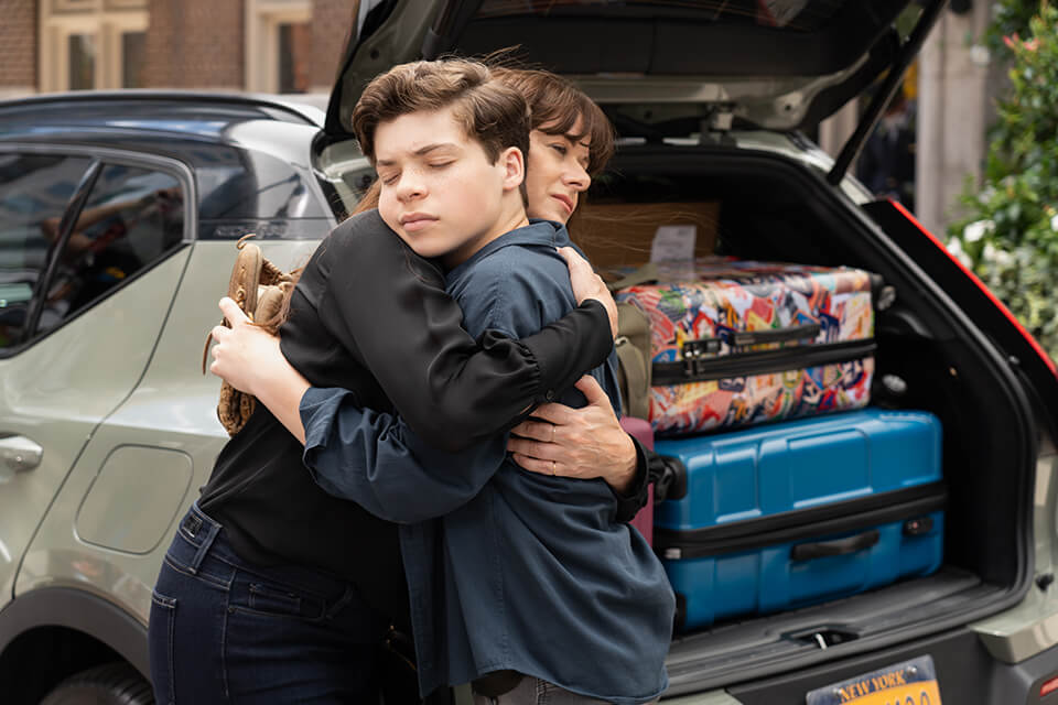 A mother hugs her son in front of a car with suitcases in the trunk
