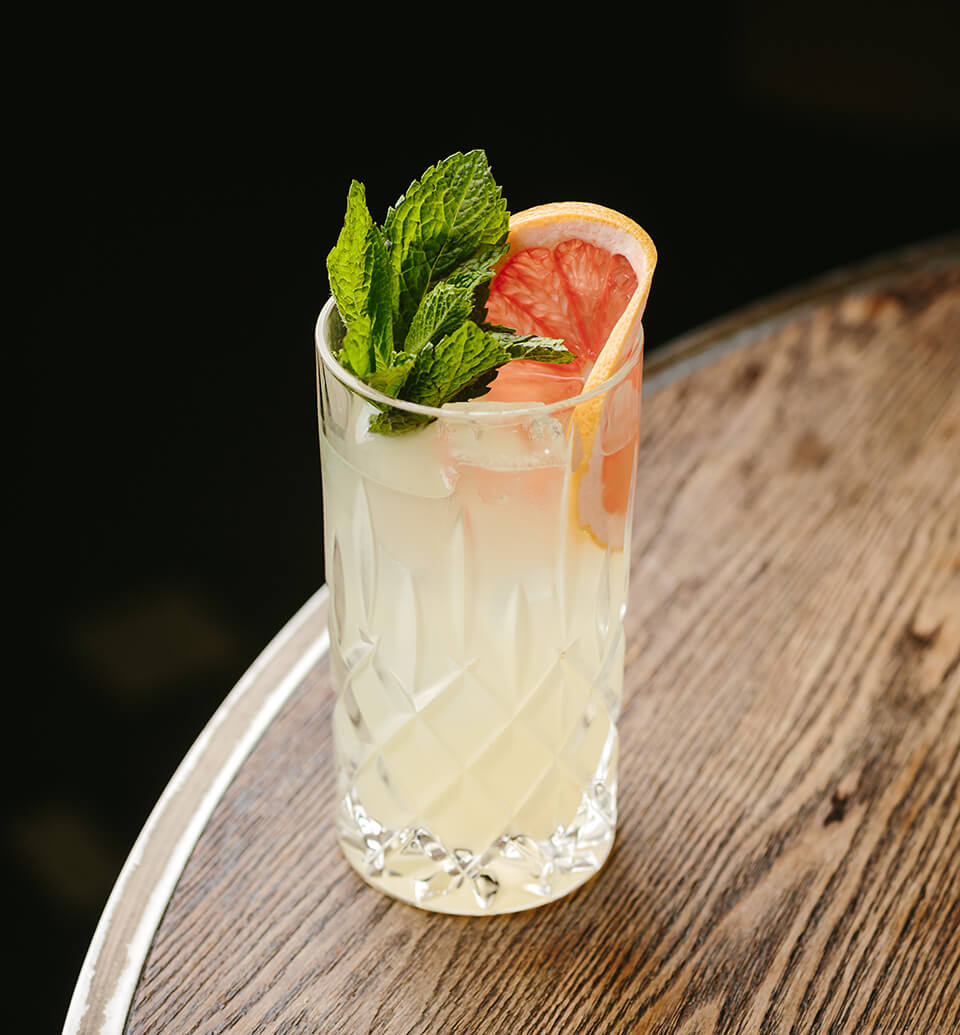 A cocktail garnished with a grapefruit wheel and mint on a wooden table against a dark background