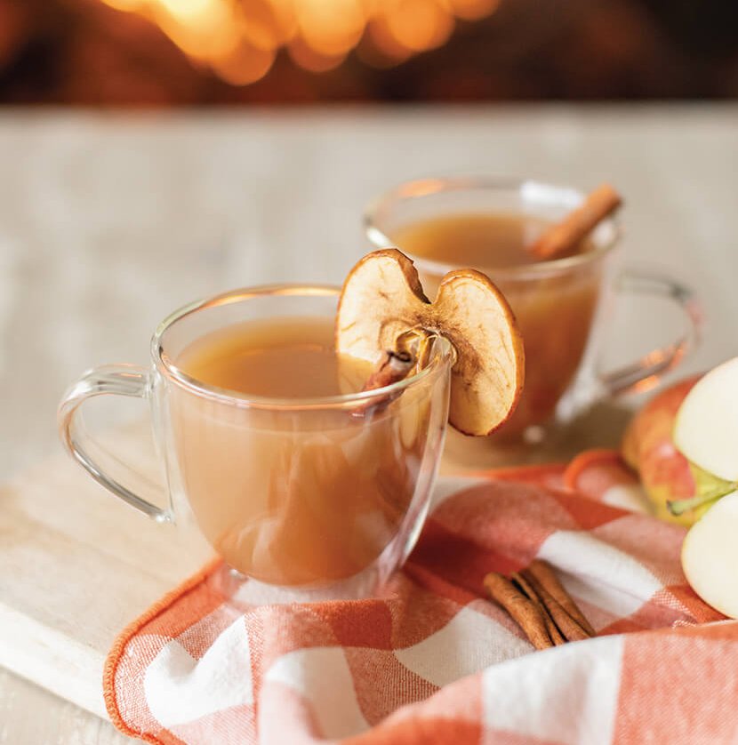 Two mugs of apple cider on a marble surface with a fire in the hearth in the background