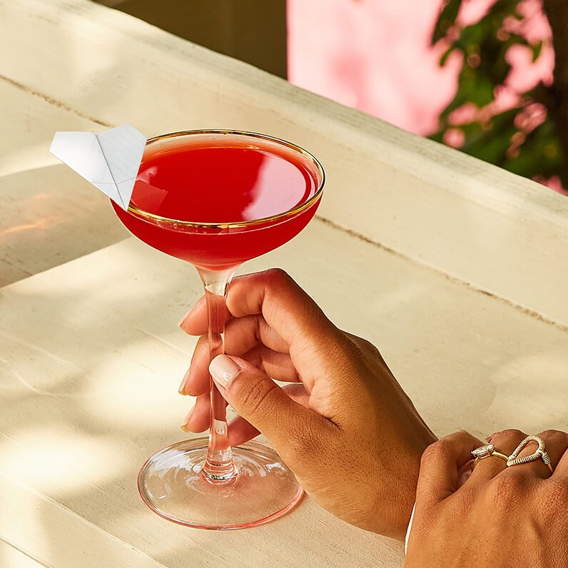 A woman's hand holding a cocktail garnished with a small paper plane.