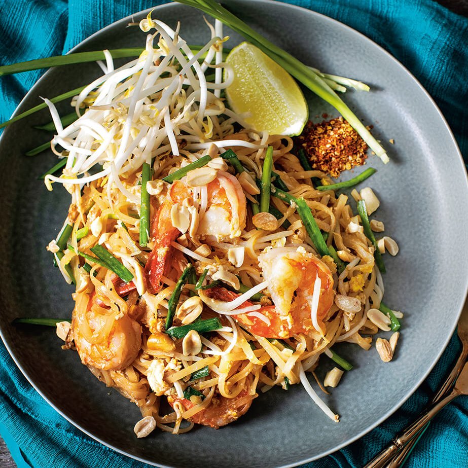 A dish of pad thai on a blue cloth with a fork and spoon lying next to it