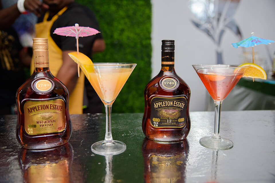 Bottles of rum and cocktails on a clear glass surface and greenery visible in the background
