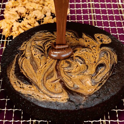 Ganache is poured over a single-layer chocolate cake on a drying rack, with cornflake crunch topping visible in the background