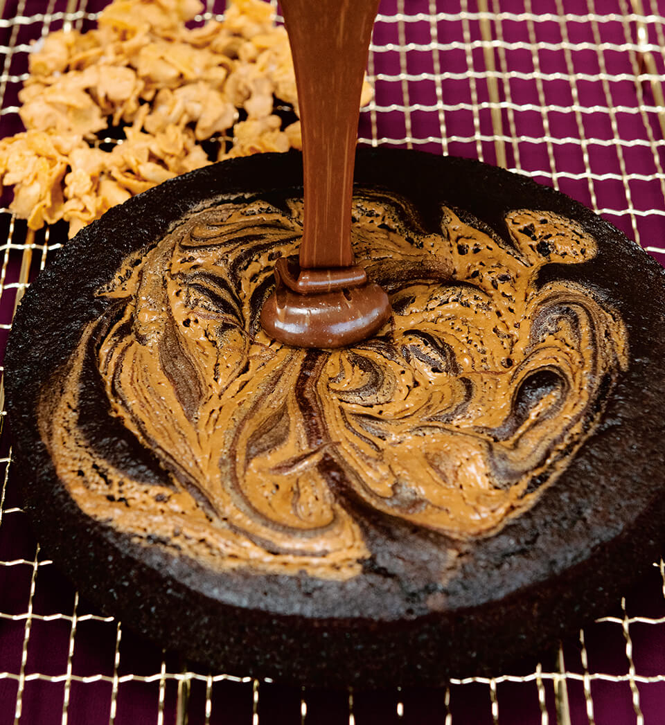 Ganache is poured over a single-layer chocolate cake on a drying rack, with cornflake crunch topping visible in the background