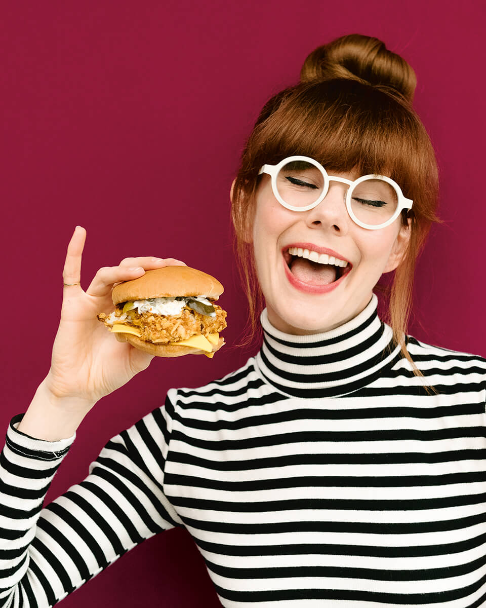 A woman, Mary Berg, holds a fried fish sandwich in front of a red background