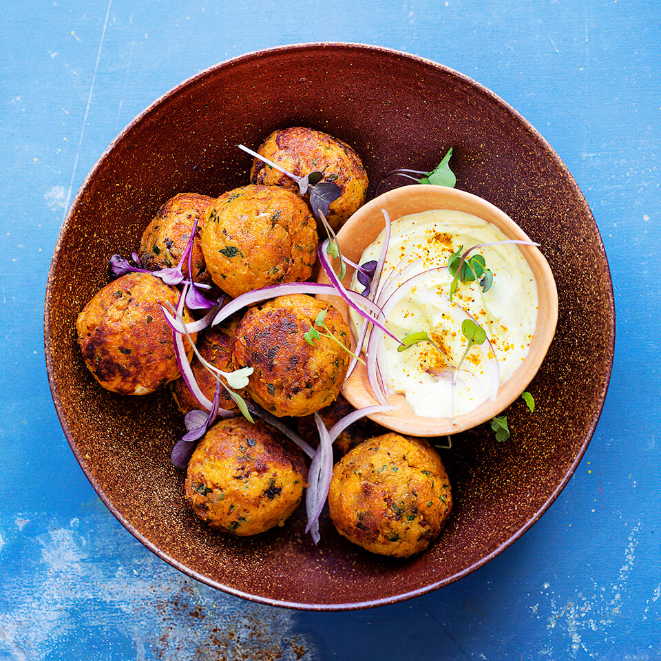 A bowl of fried lentil and potato balls with a dish of white dip on a blue surface