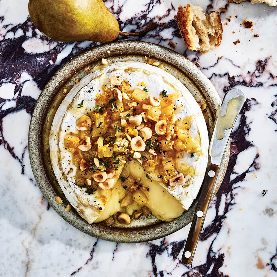 A dish of brie on a marble surface with a whole pear and a knife