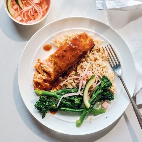 A white plate with salmon, rice and broccolini with a small dish of sauce next to it