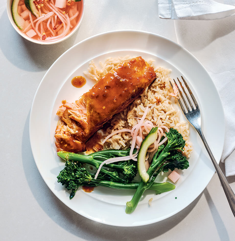 A white plate with salmon, rice and broccolini with a small dish of sauce next to it