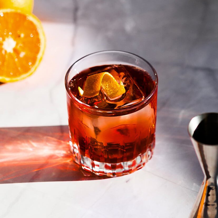 A negroni cocktail on a marble surface with a jogger and oranges next to it