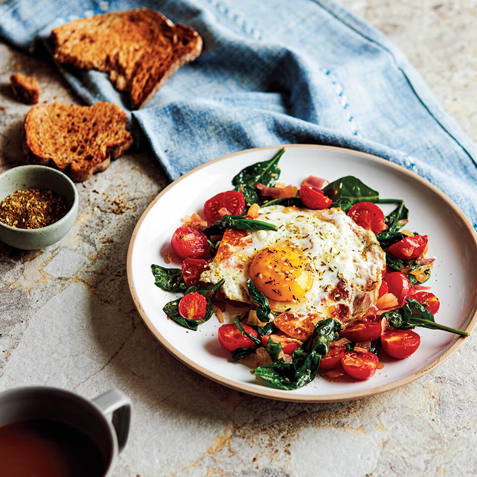 A dish with eggs, spinach, tomatoes and halloumi with toast, tea, and a light blue napkin visible in the background