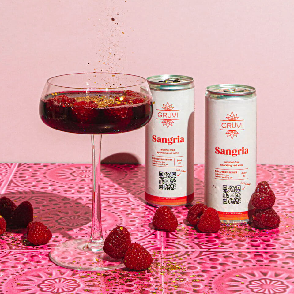 Cans and a glass of Sangria with raspberries around them on a bright pink surface