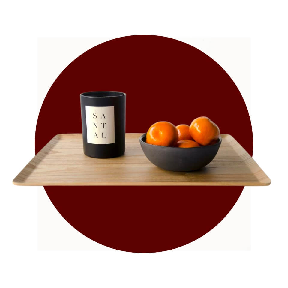 A wooden serving tray with a black jar candle and a black bowl filled with oranges over a maroon circle