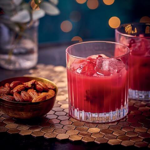 Two glasses with a red cocktail and a small dish of almonds on a wooden placemat made of hexagons