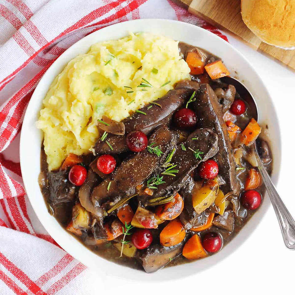 A white dish with mashed potatoes and pot roast on a white surface with a red-and-white tea towel underneath