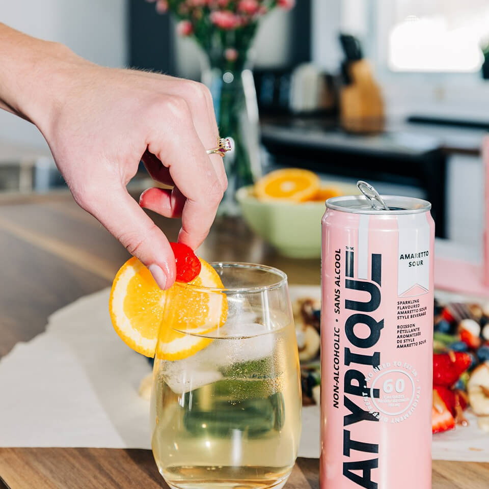 A pink can and glass with a person's hand placing a lemon wheel garnish