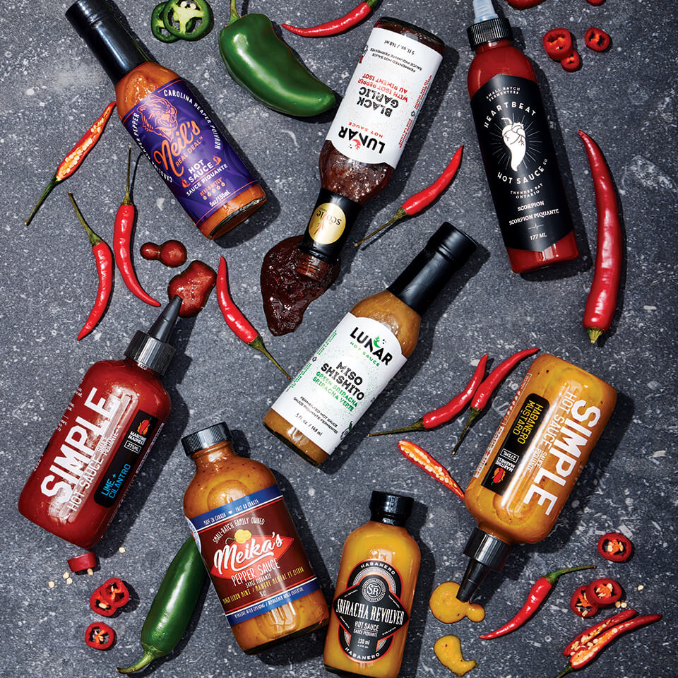 A variety of hot sauce bottles scattered amongst chili peppers on a grey surface