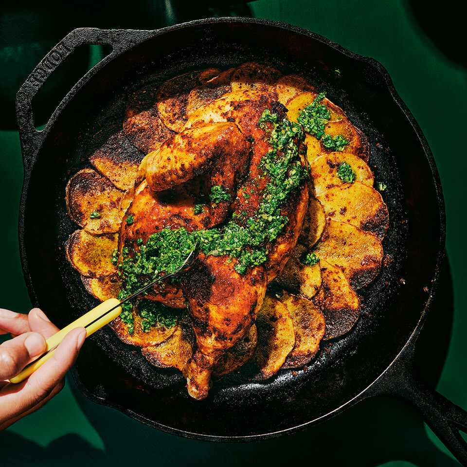 A skillet with chicken covered in green sauce and a person's hand visible in the bottom right corner