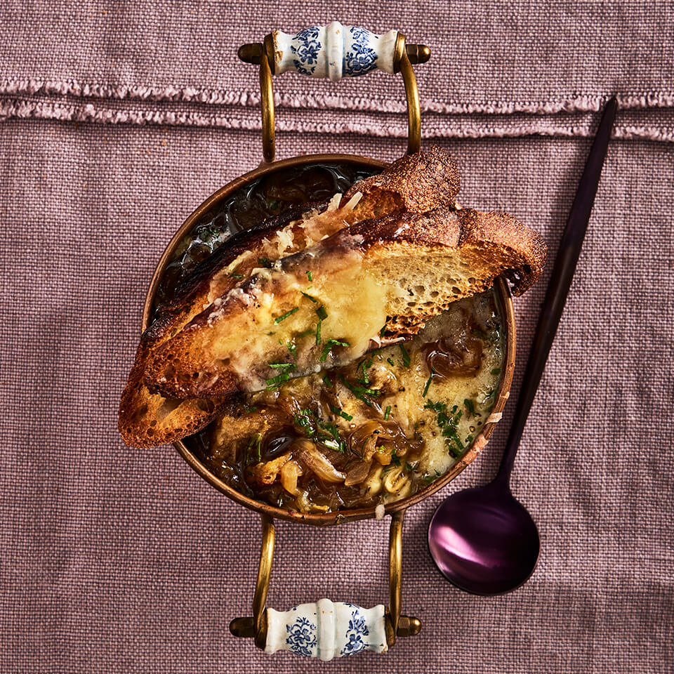 A dish of French onion soup with bread and a purple spoon on a light purple cloth