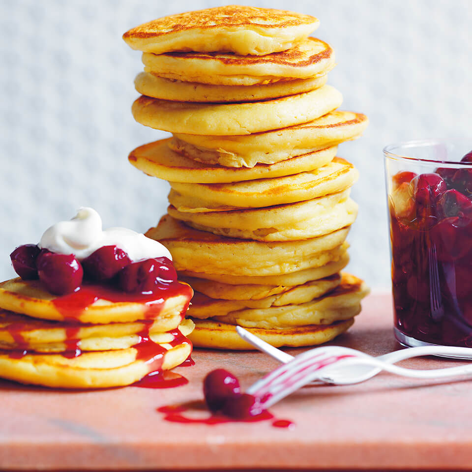 A stack of pancakes with sour cherries on a light pink surface with a white background