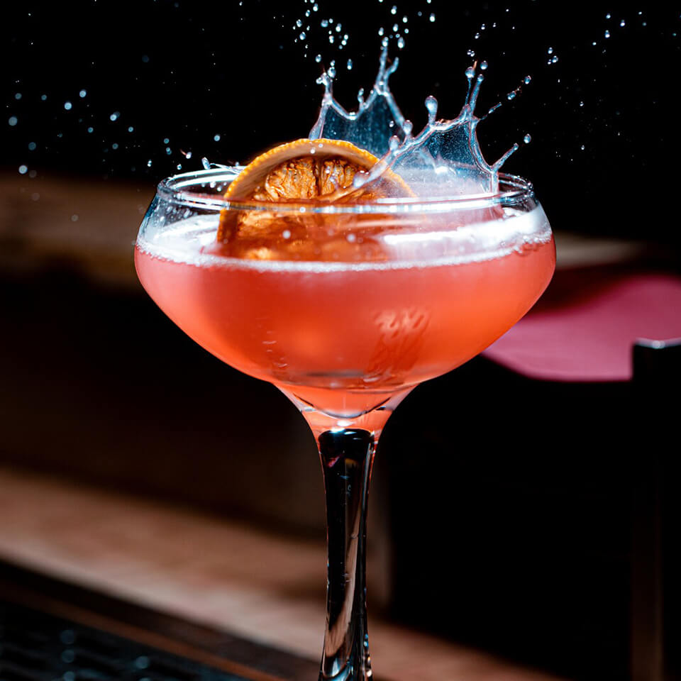 A dried lemon wheel splashing into a red cocktail in a coupe glass