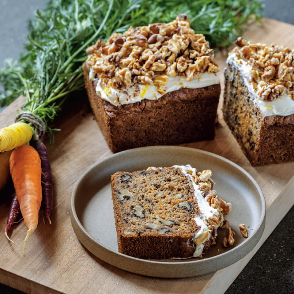 A wooden cutting board with a bushel of carrots and frosted carrot cake loaf