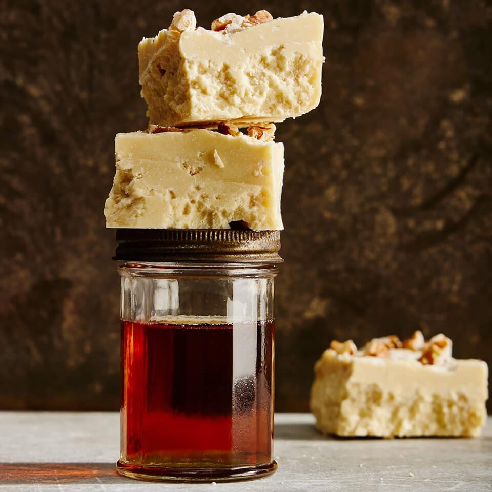 A jar of maple syrup with squares of white fudge stacked on top and one in the background.