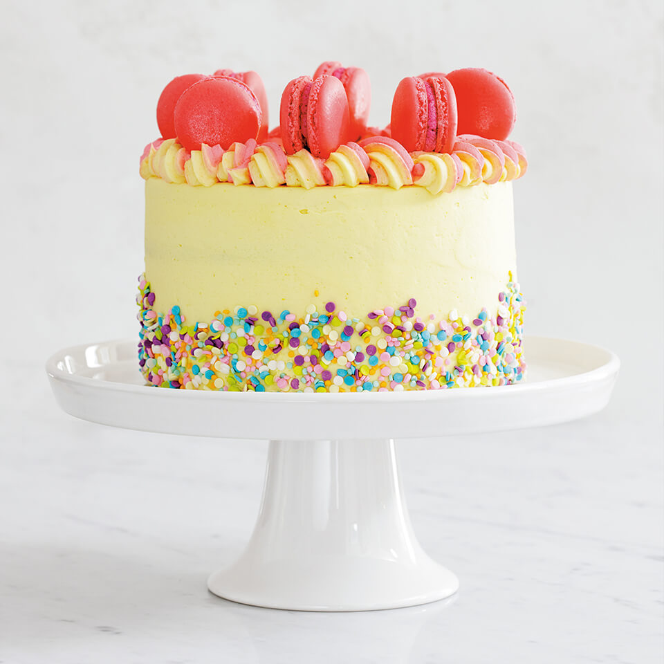 A cake frosted with light yellow and decorated with pink macarons and rainbow sprinkles on a white cake stand