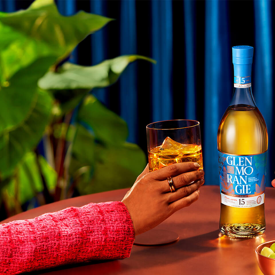 A person's hand holding a glass of whisky with the bottle and a plant visible