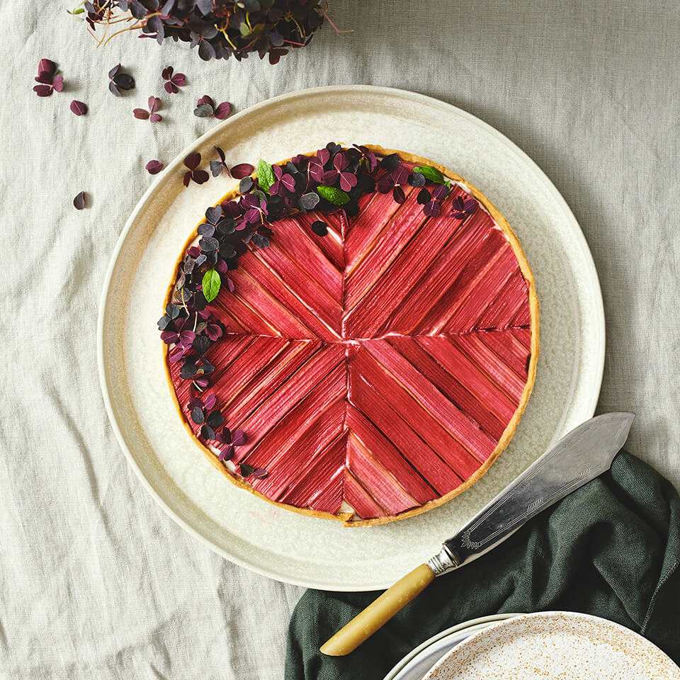 A pie with sliced rhubarb on top on a white plate with linens and a knife next to it