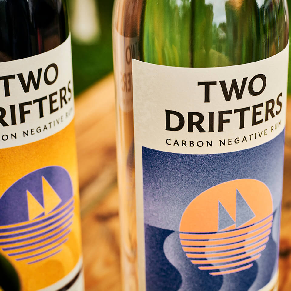 A closeup of two rum bottles, one white and one amber, with the words "Two Drifters Carbon Negative Rum"