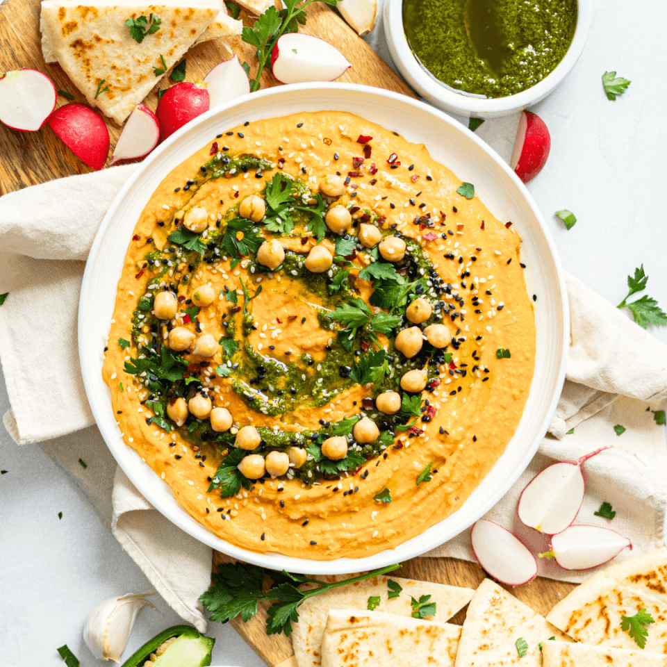 Overhead shot of bowl of carrot hummus with pita bread and vegetables.