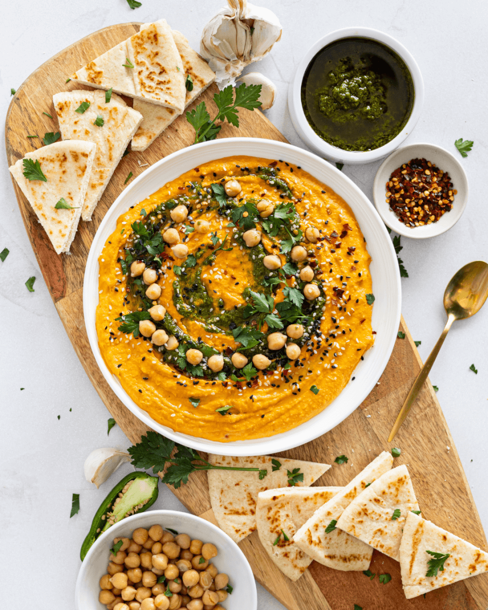 Overhead shot of bowl of carrot hummus with pita bread and vegetables.