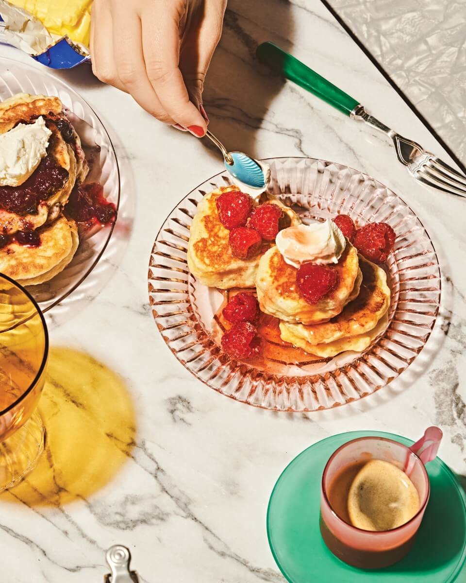 A person's hand holding a spoon over a dish with pancakes and fruit