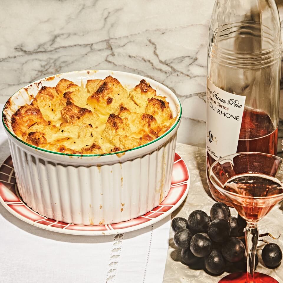 A ramekin with a cheese soufflé on a plate on a marble surface, with a bottle and partly-poured glass and grapes next to it